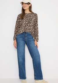 Blusa de manga larga con estampado de leopardo en beige y marrón, combinada con unos vaqueros de pierna ancha y cintura alta en azul. Zapatillas de color claro y una gorra completan el look.