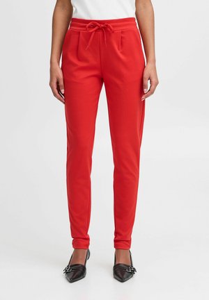 Woman wearing bright red slim-fit pants with a drawstring waist, paired with black pointed-toe buckle shoes, standing on a white background.