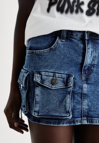 Denim mini svārki ar bālētu zilu krāsu, divām lielām priekšējām kabatām un metāla pogu akcents. Gluda tekstūra ar redzamām šuvēm.