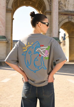 Woman with hair in a bun wearing sunglasses and a gray shirt featuring a colorful shark graphic biting a pair of legs, standing outdoors near arches.