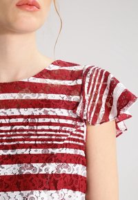 Blouse en dentelle présentant un motif à rayures rouges et blanches, manches à volants et détails floraux, mettant en valeur une texture transparente et un design délicat.