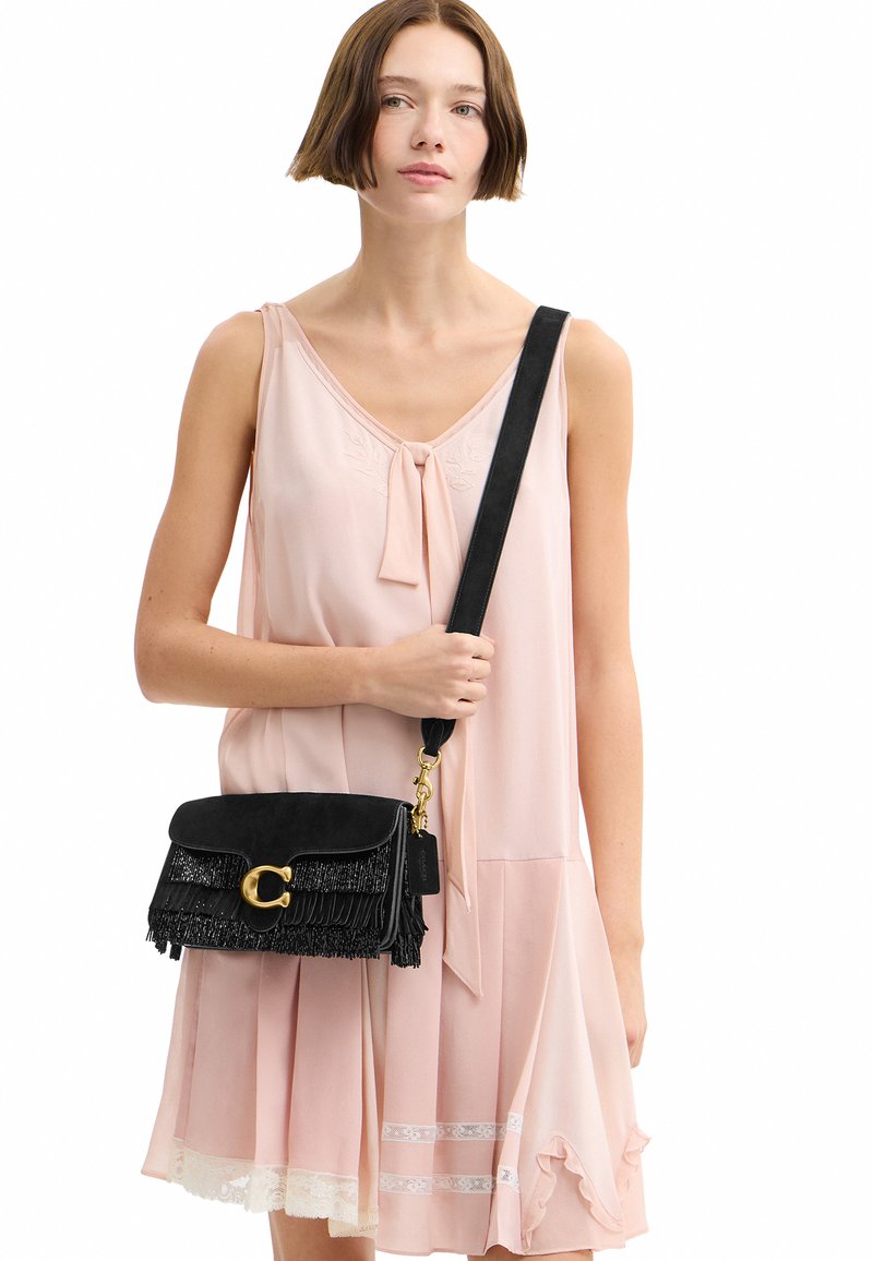 Femme en robe rose clair sans manches tenant un sac bandoulière noir à franges avec une large sangle et une fermeture dorée.