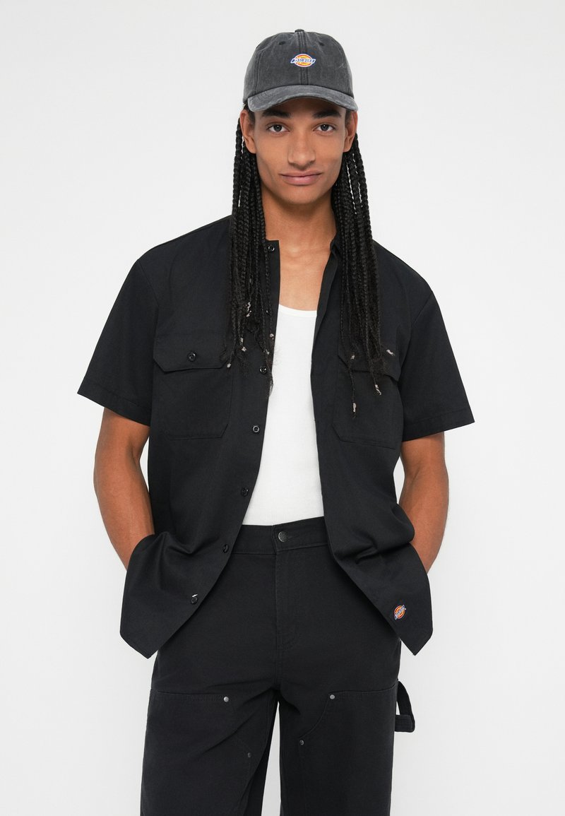 Chemise noire à manches courtes avec deux poches poitrine, associée à un pantalon sombre. Casquette grise avec logo. Couleur unie et design décontracté.