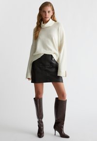 Maglione a collo alto crema con maniche a coste, gonna mini in pelle nera con tasche frontali e stivali cuissard in pelle stampa cocco marroni con tacco a spillo.