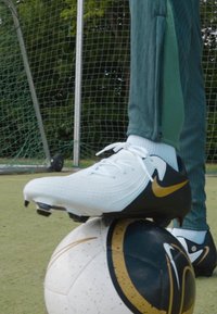 Vita och svarta Nike fotbollsskor med guldaccenter som vilar på en svart- och vitmönstrad fotboll, placerad på en grön gräsyta.