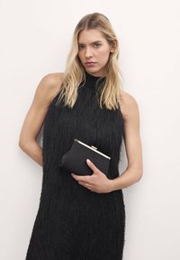 Abito nero con frange e collo alto; la modella tiene una piccola pochette nera con chiusura dorata. La texture liscia contrasta con il design a frange.