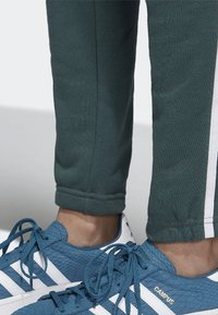 Dark green athletic joggers with elastic cuffs, paired with blue textured sneakers featuring white stripes and branding on the side.