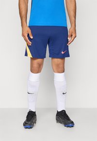 Mees sportlane kannab siniseid ja kollaseid spordilühikesi pükse, sinist särki, valgeid põlvikuid Nike ja musti jalatseid, seistes hallil põrandal.