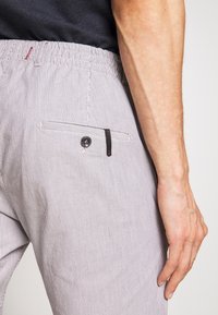 Muž v světle šedých pruhovaných kalhotách s knoflíkovou zadní kapsou a černým elastickým pasem, stojí s uvolněnou pravou rukou.