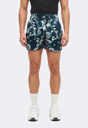 Mann trägt dunkelblaue, floral gemusterte Champion-Shorts, weiße Socken und weiße Sneaker, steht mit entspannten Armen.