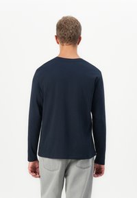 Man med kort lockigt hår som bär en marinblå långärmad skjorta och ljusgrå jeans, står vänd bort mot en enfärgad vit bakgrund.