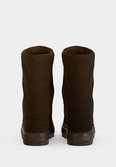Paire de bottes hautes en daim marron foncé avec semelles en caoutchouc, vues de dos sur un fond clair uni.