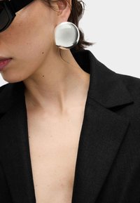 Silberne, kreisförmige Ohrringe mit glänzender Oberfläche, getragen zu einem schwarzen Blazer. Die Ohrringe sind groß und glatt und reflektieren das Licht.