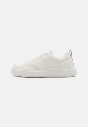 Des sneakers blanches avec un dessus en cuir lisse, un bout rond, une semelle plate et un design minimaliste, agrémentées d'une languette grise au talon.