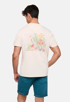 Hombre con camiseta beige con estampado tropical colorido de flores, hojas, sandía y un velero, combinado con pantalones cortos azules remangados.