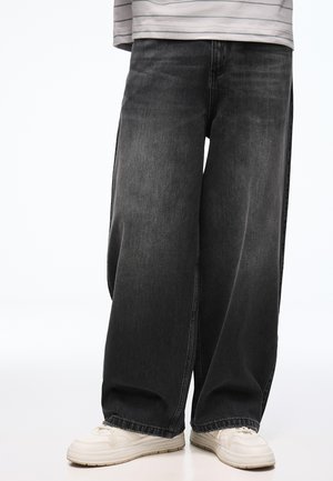 Person trägt lockere schwarze Denim-Jeans und weiße Sneakers, steht vor einem einfarbigen hellen Hintergrund.