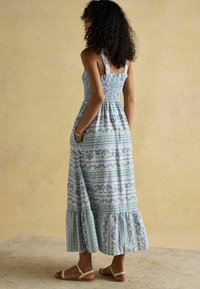 Maxi šaty v modro-bielej farbe s kvetinovými a pruhovanými vzormi, smockovaným živôtikom, širokými ramienkami a rozšíreným lemom. Nosené s ľahkými sandálmi.