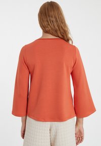 Kupferfarbener Oberteil mit weitem, ausladendem langen Ärmel und rundem Halsausschnitt. Aus einem glatten, weichen Stoff gefertigt. Schlichtes, minimalistisches Design.