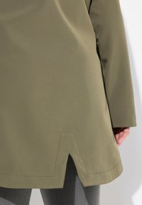 Cappotto di tessuto verde oliva con taglio dritto, caratterizzato da un dettaglio a forma di V all'orlo e cuciture visibili. Design casual con maniche lunghe.