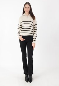 Cremfarbener, gerippter Pullover mit schwarzen Streifen, Reißverschlusskragen, kombiniert mit schwarzen Schlaghosen und spitzen schwarzen High Heels. Einfaches Design, strukturierter Stoff.