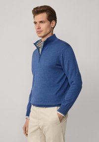 Pull bleu avec un col à zip quart, manches longues, fabriqué en tissu tricoté texturé, avec un intérieur rayé au niveau du col.