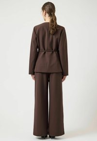 Bruine op maat gemaakte blazer met riem, gecombineerd met brede bruine broek. Soepele textuur, minimalistisch ontwerp en een strak, gestructureerd silhouet.