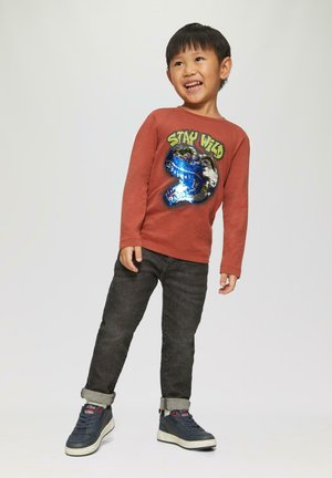 Langekleurige shirt in roestkleur met de tekst "STAY WILD" en een paillettengraphic, gecombineerd met donkere denim jeans en sneakers.