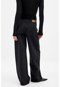 Black denim wide-leg jeans with a textured pattern and visible stitching. Features a brown label at the back waistband. Plain smooth fabric.