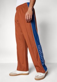 Bruine joggingbroek met blauwe zijkanten en witte bies, en een prominent logo. Het materiaal lijkt zacht en heeft een losse fit.