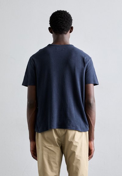 T-shirt en coton bleu marine à manches courtes, col rond, coupe décontractée et texture subtile ; porté avec un pantalon beige.