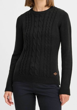 Femme portant un pull en maille torsadée noir à manches longues et un pantalon sombre, debout devant un fond blanc uni.