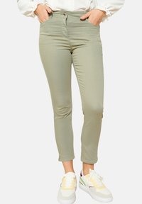 Zelene slim-fit hlače z gladko teksturo, petimi žepi in gumbom za zapiranje. Kombinirane z belimi in pastelno obarvanimi športnimi copati.