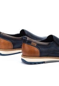 Zapatos de cuero azul oscuro con detalles en cuero marrón, diseño tipo slip-on y suela blanca con detalles en azul. Textura suave y costuras visibles.