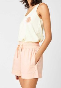 Kobieta w bezrękawniku w kolorze kremowym z okrągłym pomarańczowym logo oraz jasnoróżowych szortach na elastycznej talii z troczkami, z rękami w kieszeniach.