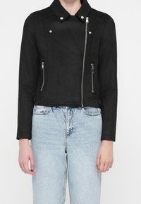 Giacca cropped in faux suede nera con chiusura a zip, colletto a scatto e due tasche frontali con zip. Texture morbida e design minimalista.