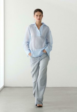 Kobieta idąca w pomieszczeniu, ubrana w przezroczystą jasnoniebieską bluzkę, jasnoniebieskie dżinsy i czarne sandały, z rękami w kieszeniach, na tle białej ściany.