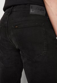 Los vaqueros de mezclilla negra presentan una textura suave, un bolsillo trasero con costura sutil y una etiqueta de cuero negro con la marca en la pretina.