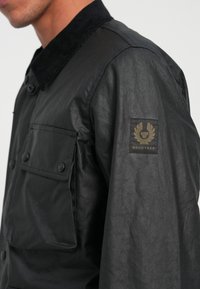 Black jacket with a smooth finish, featuring a corduroy collar, front pocket with buttons, and a Belstaff logo patch on the sleeve.