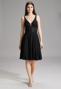 Robe noire avec un corsage ajusté en sequins et une jupe évasée et fluide. Les bretelles ajustables ajoutent un détail. Le mannequin porte des talons argentés à lanières.
