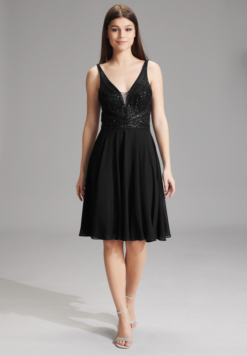 Robe noire avec un corsage ajusté en sequins et une jupe évasée et fluide. Les bretelles ajustables ajoutent un détail. Le mannequin porte des talons argentés à lanières.