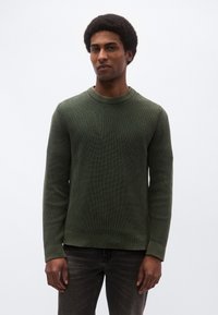 Zeleni pulover iz rebrastega platna z okroglim izrezom, dolgimi rokavi in teksturirano površino, kombiniran s temno sivimi kavbojkami.