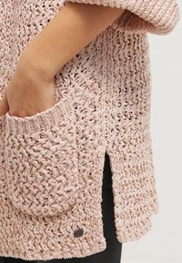 Ręka w kieszeni luźno plecionego, jasnoróżowego swetra z bocznym rozcięciem, noszonego na czarnych spodniach.