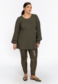 Olivgrüne metallische Strick-Tunika mit Glockenärmeln, kombiniert mit glänzenden olivfarbenen Leggings und flachen Slippers mit Verzierungen.