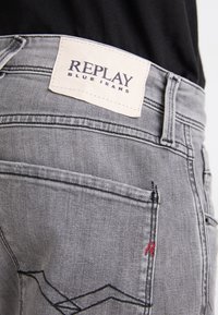 Jeans de denim gris con una etiqueta de cuero con la inscripción "REPLAY BLUE JEANS" en relieve. Detalle notable de costura roja y diseño estándar de bolsillo.