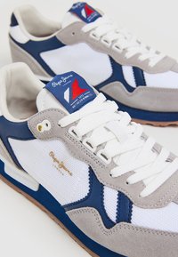 Sneaker grigi, bianchi e blu navy realizzati in materiali a rete e scamosciati. Presentano lacci bianchi, una suola testurizzata e un logo sulla linguetta.