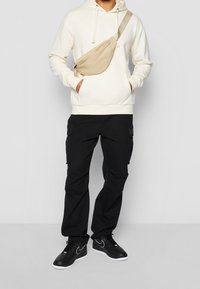 Béžový ledvinka nošená přes tělo přes krémovou mikinu; černé cargo kalhoty s bočními kapsami; černé tenisky s bílou podrážkou a logem Nike.