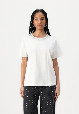 T-shirt en coton blanc avec un col rond orné d'accents argentés. Associé à un jean foncé orné de clous métalliques.