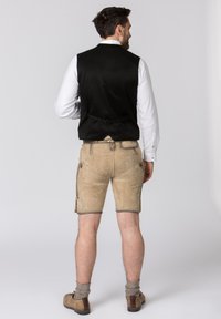 Schwarze Satinweste über einem weißen Hemd, kombiniert mit hellbraunen Leder-Shorts mit Seitentaschen und grauen Bündchen, getragen mit braunen Schuhen.