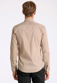 Camicia beige a maniche lunghe con colletto, polsini con bottoni e texture liscia. Presenta un orlo curvo e una leggera lucentezza.