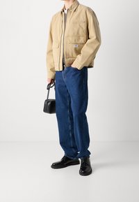 Beżowa bawełniana kurtka bomberka z zamkiem i kieszeniami, w połączeniu z niebieskimi dżinsami i czarnymi skórzanymi butami, trzymająca małą czarną torbę.